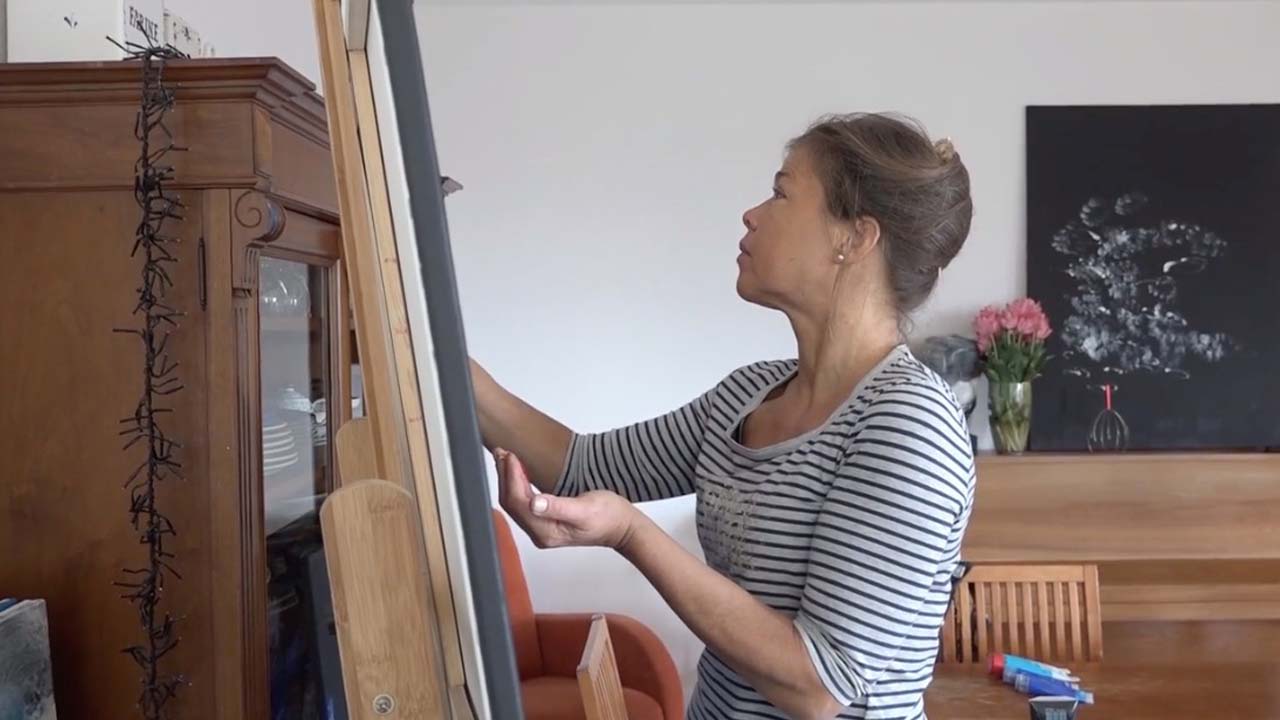 The Artist. Photo of Laura Meddens working at her easel while the painting "Tango" sits on top of a piano in the background.