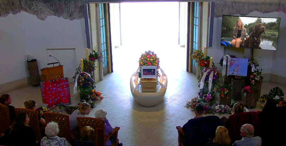 Stillscreen from streaming video of the funeral service for Laura Meddens. The coffin lays on a light colored rounded stone slab in front of tall doors open to outside, flanked by iron candle stands draped and flanked by flowers. A TV screen shows Laura riding in a canal boat with her guide dog Nugget, who lays on the floor near the speakers lectern on the left. Curved rows of seats to the rear of the frame contain family members and friends.
